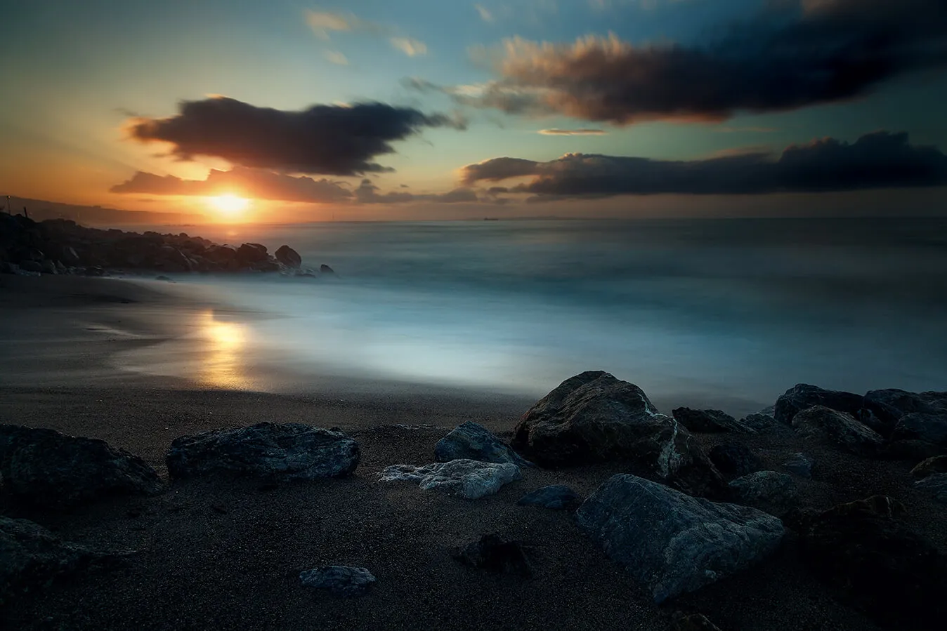 Spiaggia con rocce in primo piano, mare al tramonto in lunga esposizione.