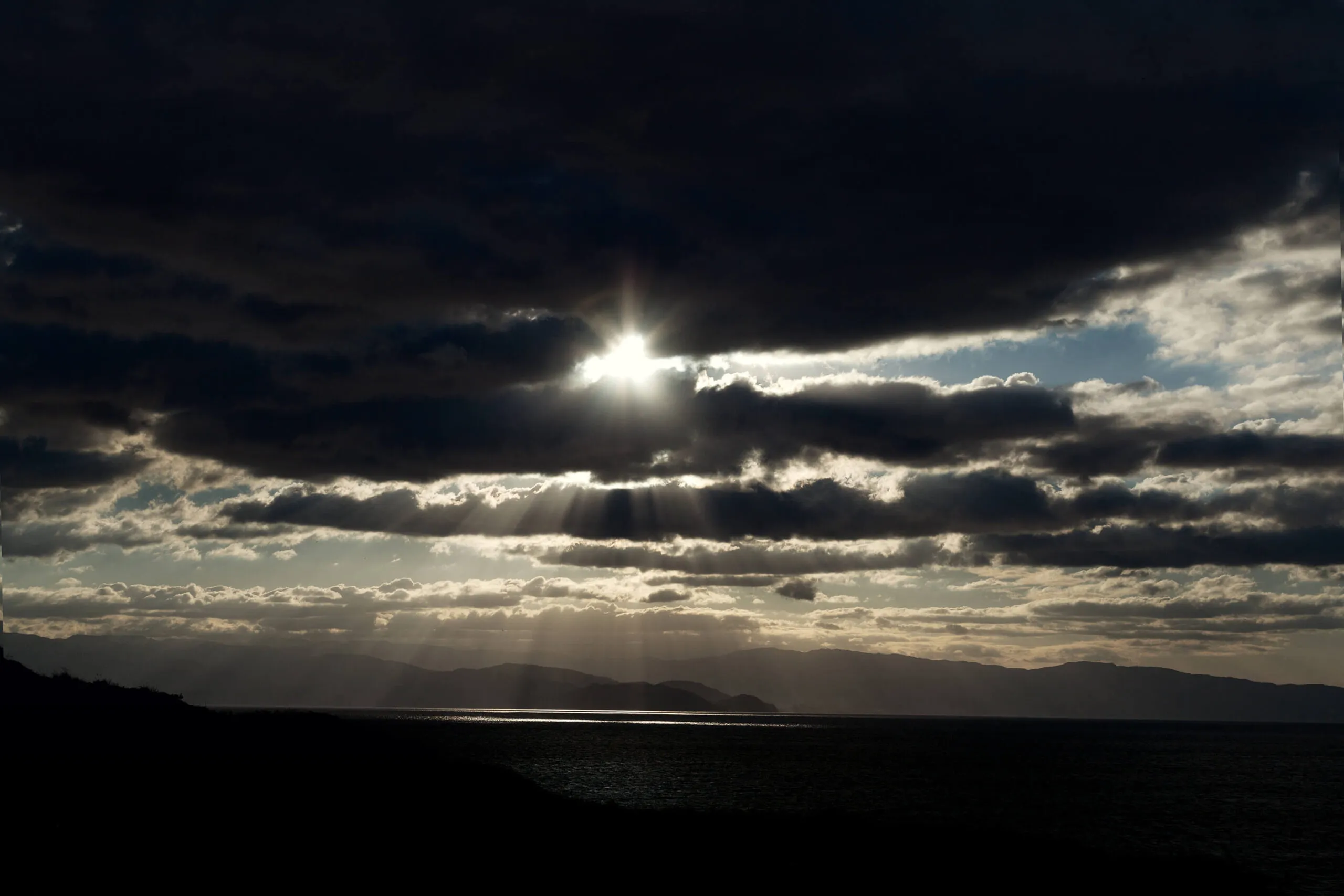 Raggi di sole tra le nuvole su colline e mare in controluce.