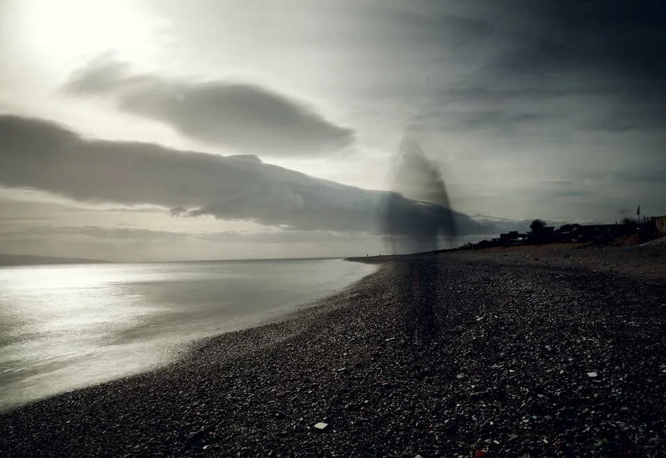 Spiaggia di ciottoli in lunga esposizione, sagoma umana sfocata, paese sullo sfondo.